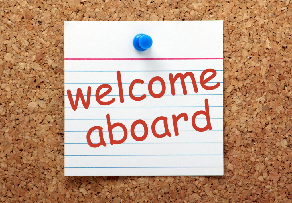 The phrase Welcome Aboard printed on a lined index card and pinned to a cork notice board